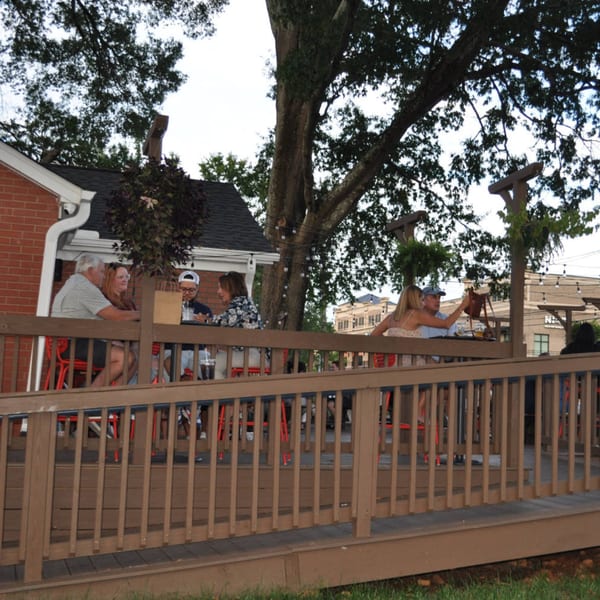 Patrons enjoy drinks and conversation on the outdoor wooden deck of Panzu Brewery under a large tree.
