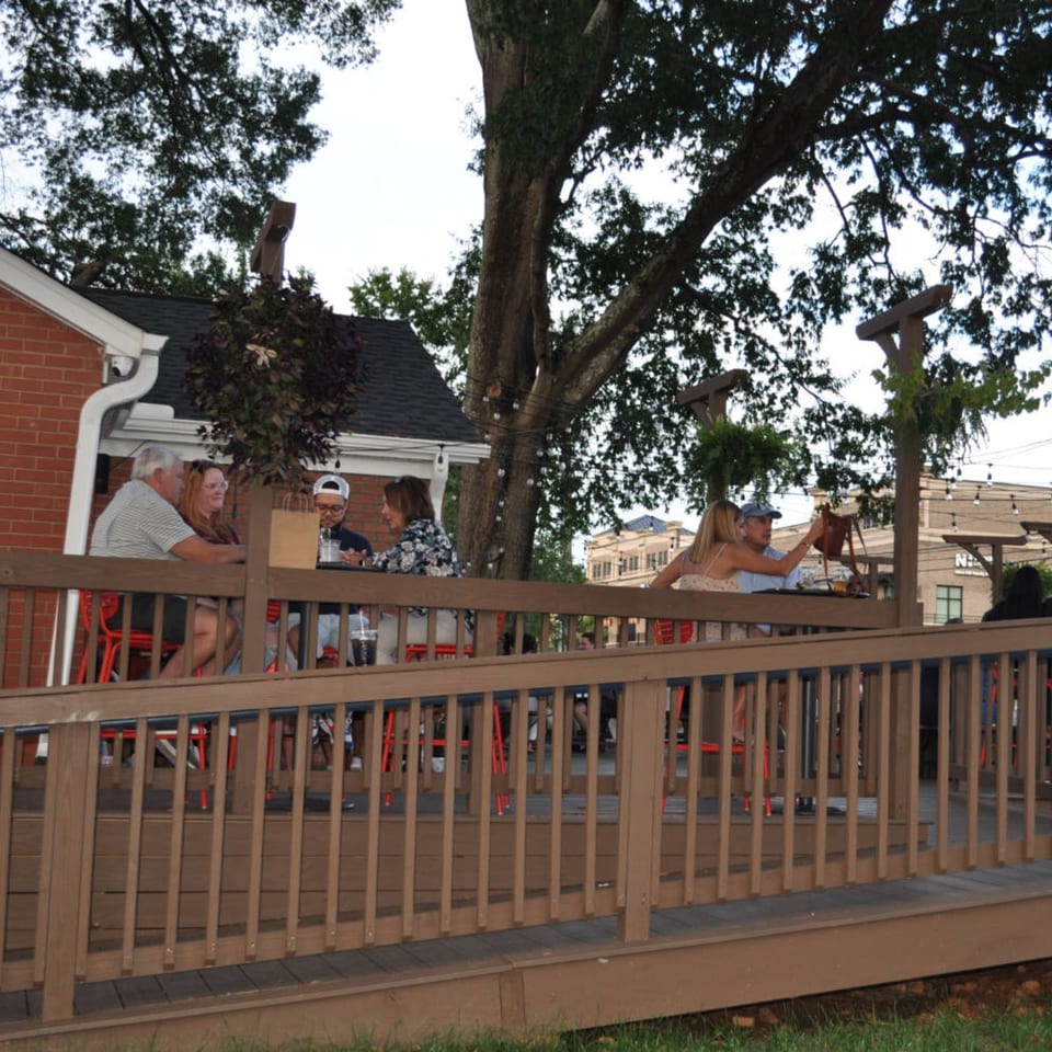 Patrons enjoy drinks and conversation on the outdoor wooden deck of Panzu Brewery under a large tree.
