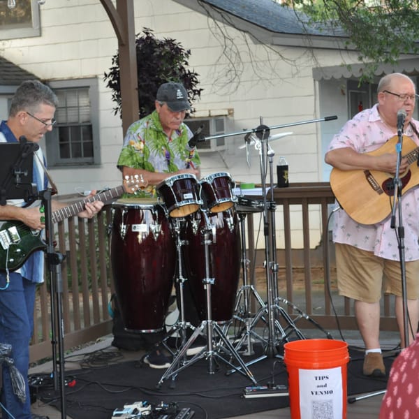 Three men performing live music outdoors at Panzu Brewery, with guitars, conga drums, and microphones.