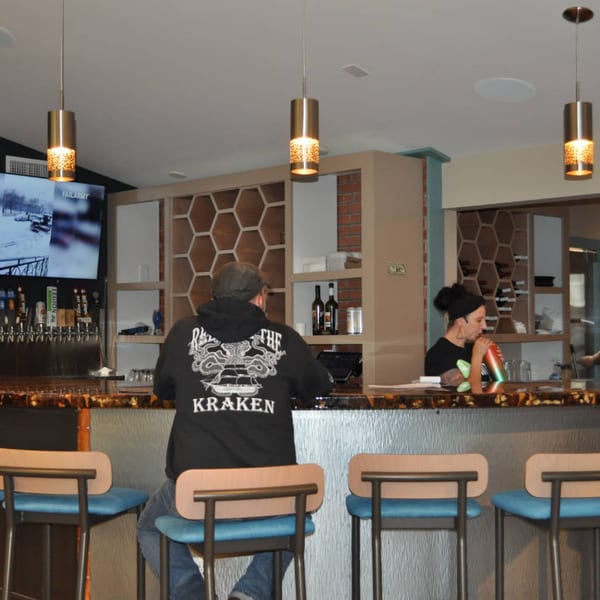 A man in a black hoodie sits at a bar with a bartender and beer taps in the background.