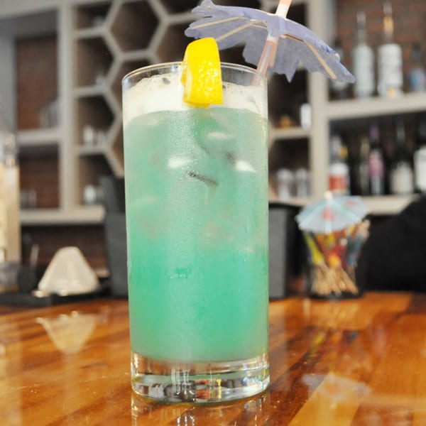 A vibrant blue cocktail with ice, a lemon wedge, and a paper umbrella on a wooden bar.