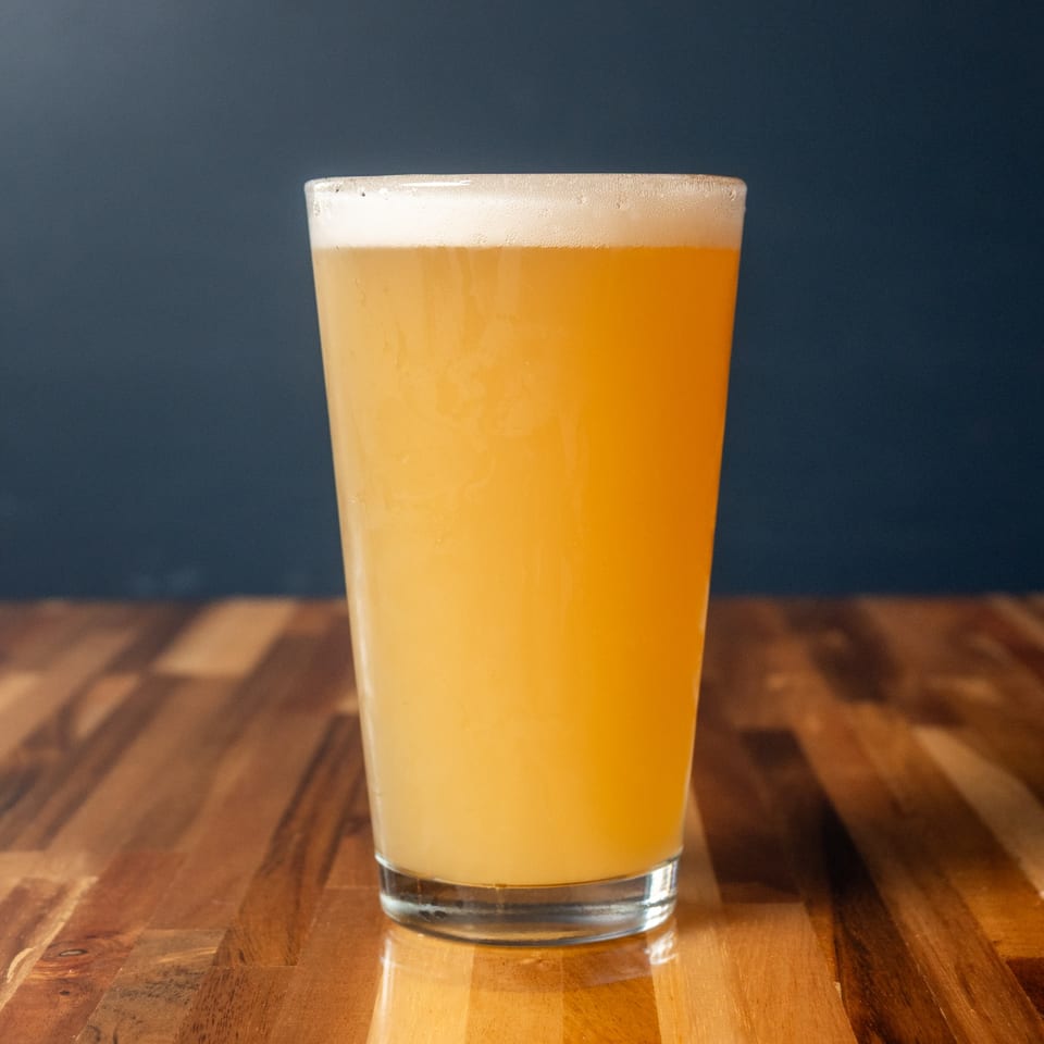 A tall glass of Panzu Brewery's hazy pale ale with a foamy head on a wooden table.