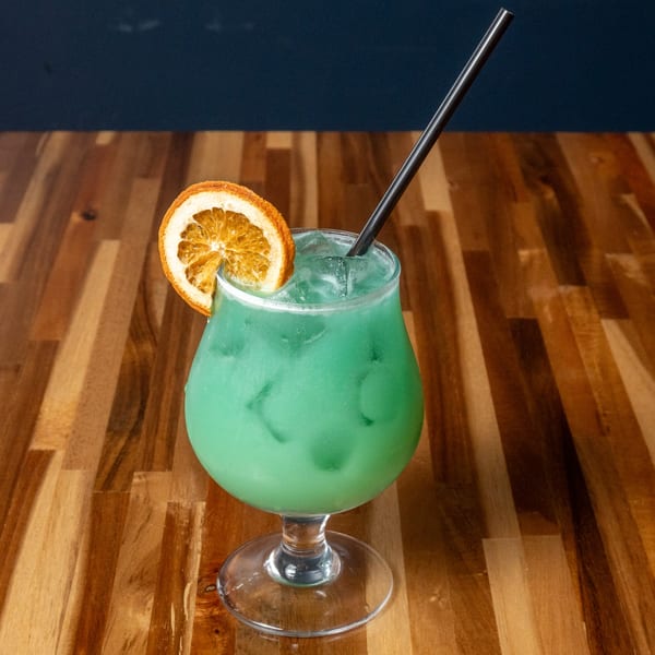 A vibrant green cocktail with ice, garnished with a dried orange slice and black straw.