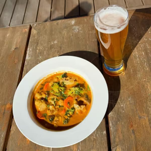 A bowl of curry with chicken and vegetables next to a glass of beer on a wooden table.