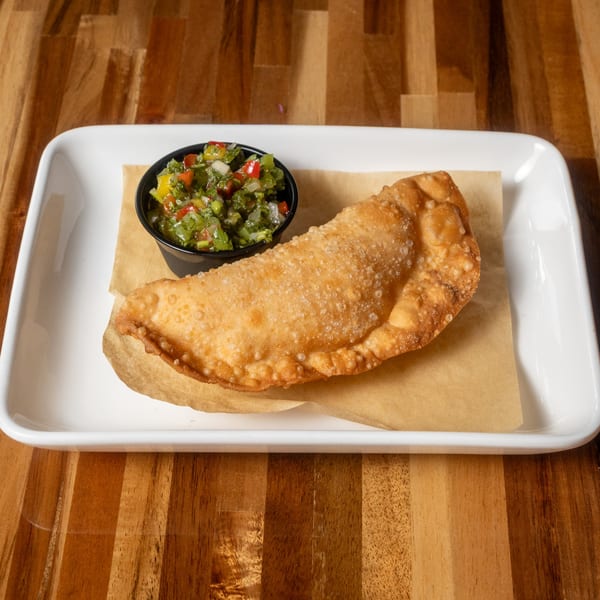A golden-brown empanada with a side of fresh salsa on a white plate at Panzu Brewery.
